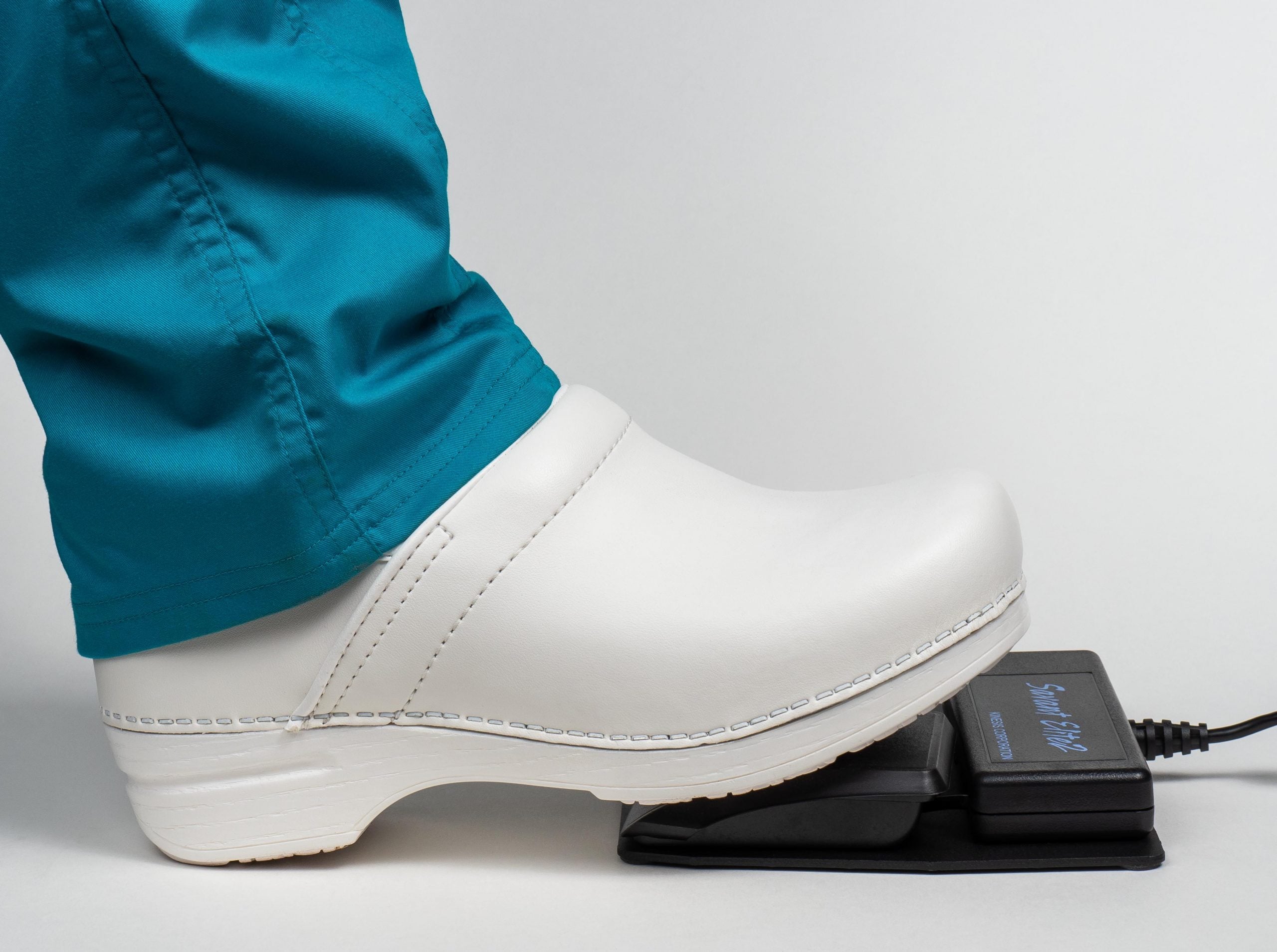 White shoe pressing electronic foot pedal, person wearing teal pants, neutral background.