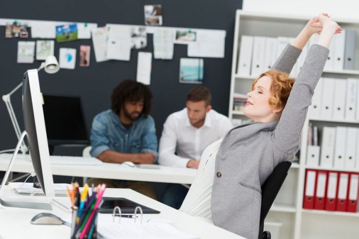 Woman stretching at desk in busy office environment, while two colleagues work in the background.