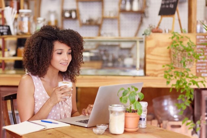 Woman enjoying coffee while working on a laptop in a cozy cafe with plants and notebooks.