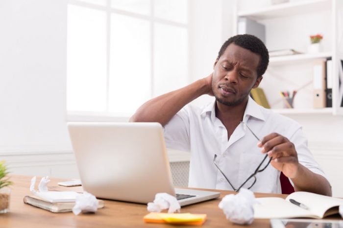Man sitting at desk with laptop, rubbing sore neck, looking tired.