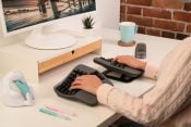 Person using ergonomic keyboard at modern desk with computer, plant, and coffee mug.
