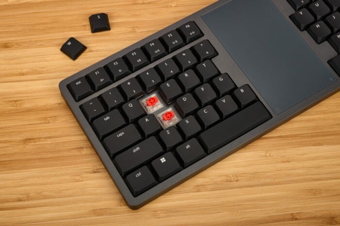 Mechanical keyboard with removed keycaps showing red switches on wooden desk.