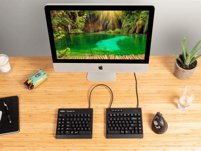 Ergonomic split keyboard on a wooden desk with a computer, mouse, notebook, and potted plant.