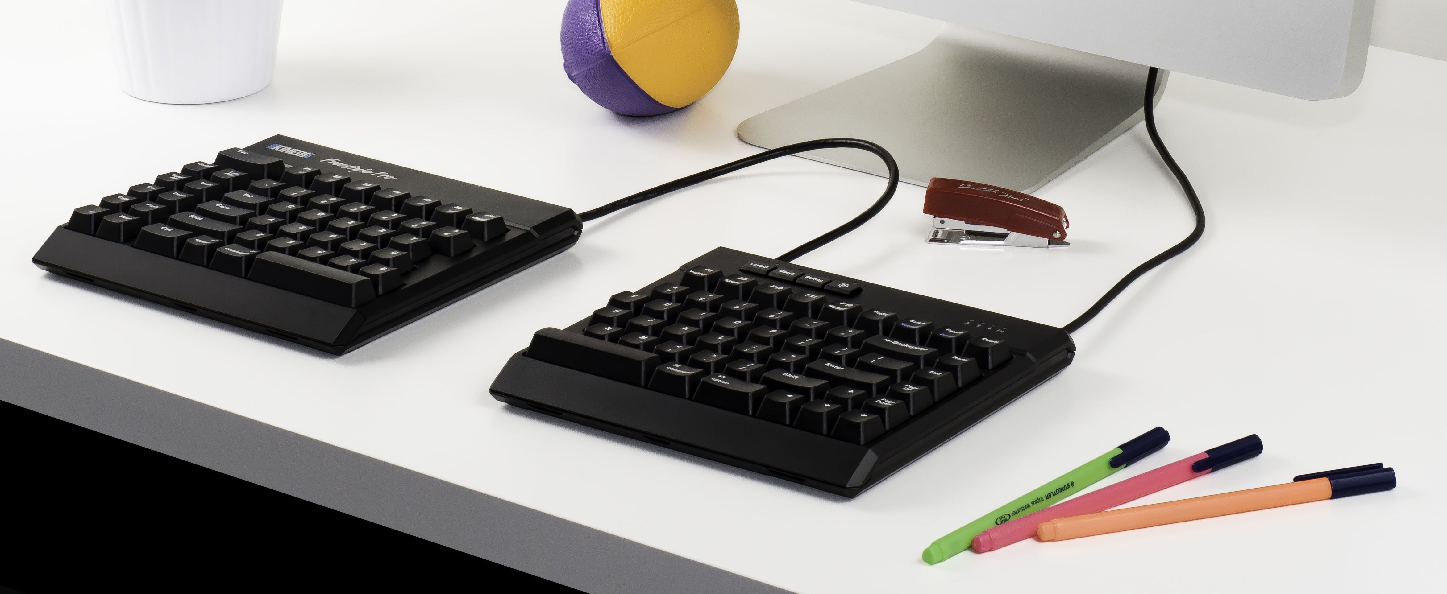 Split keyboard on desk with monitor, colorful pens, stress ball, and stapler for a modern workspace setup.