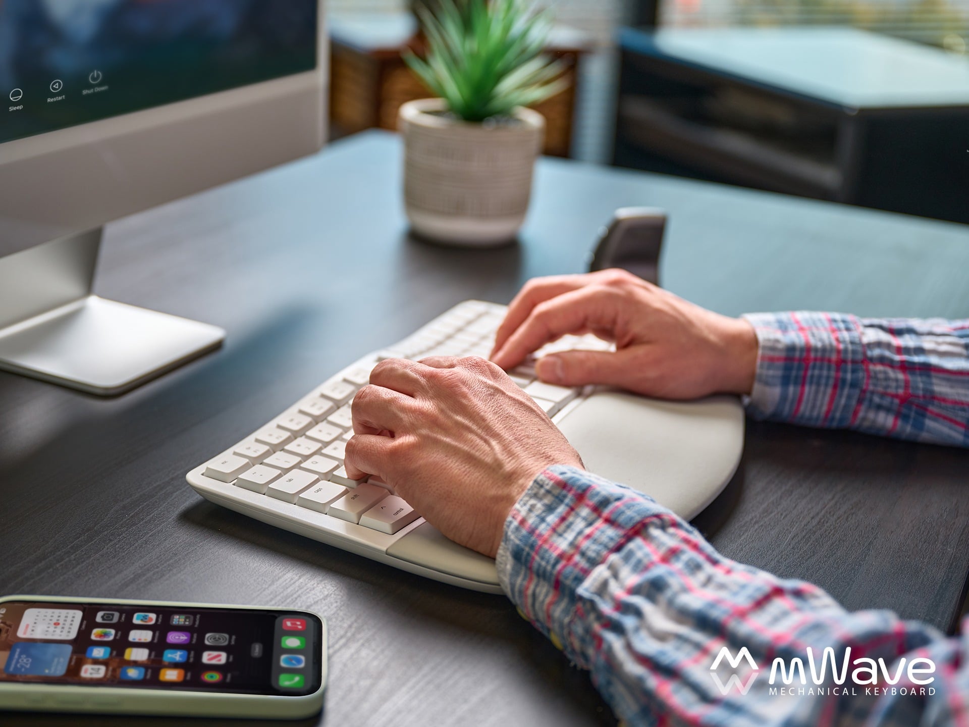 Person typing on a white keyboard at a modern desk with a smartphone and monitor nearby.