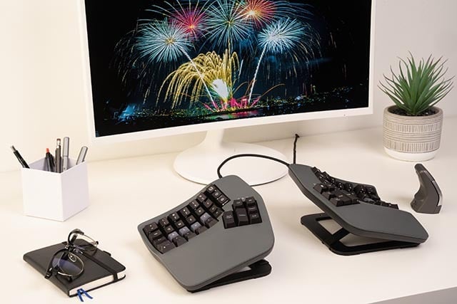 Ergonomic split keyboard setup on desk, with fireworks displayed on a monitor and a potted plant beside it.