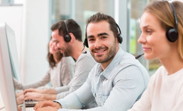 Smiling customer support team working at computers with headsets in an office setting.
