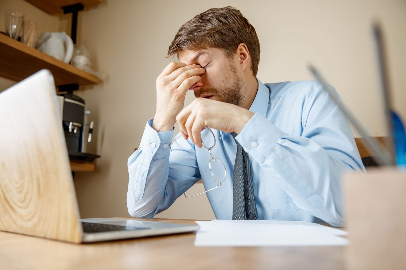 Man in office feeling stressed, holding glasses, sitting by a laptop.