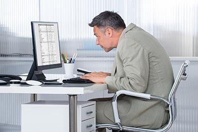 Man in suit hunched over computer at desk, focused on screen. Office setting with pens and notes visible.