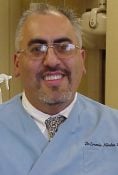 Smiling dentist in blue coat and glasses, standing inside a dental clinic with equipment in the background.