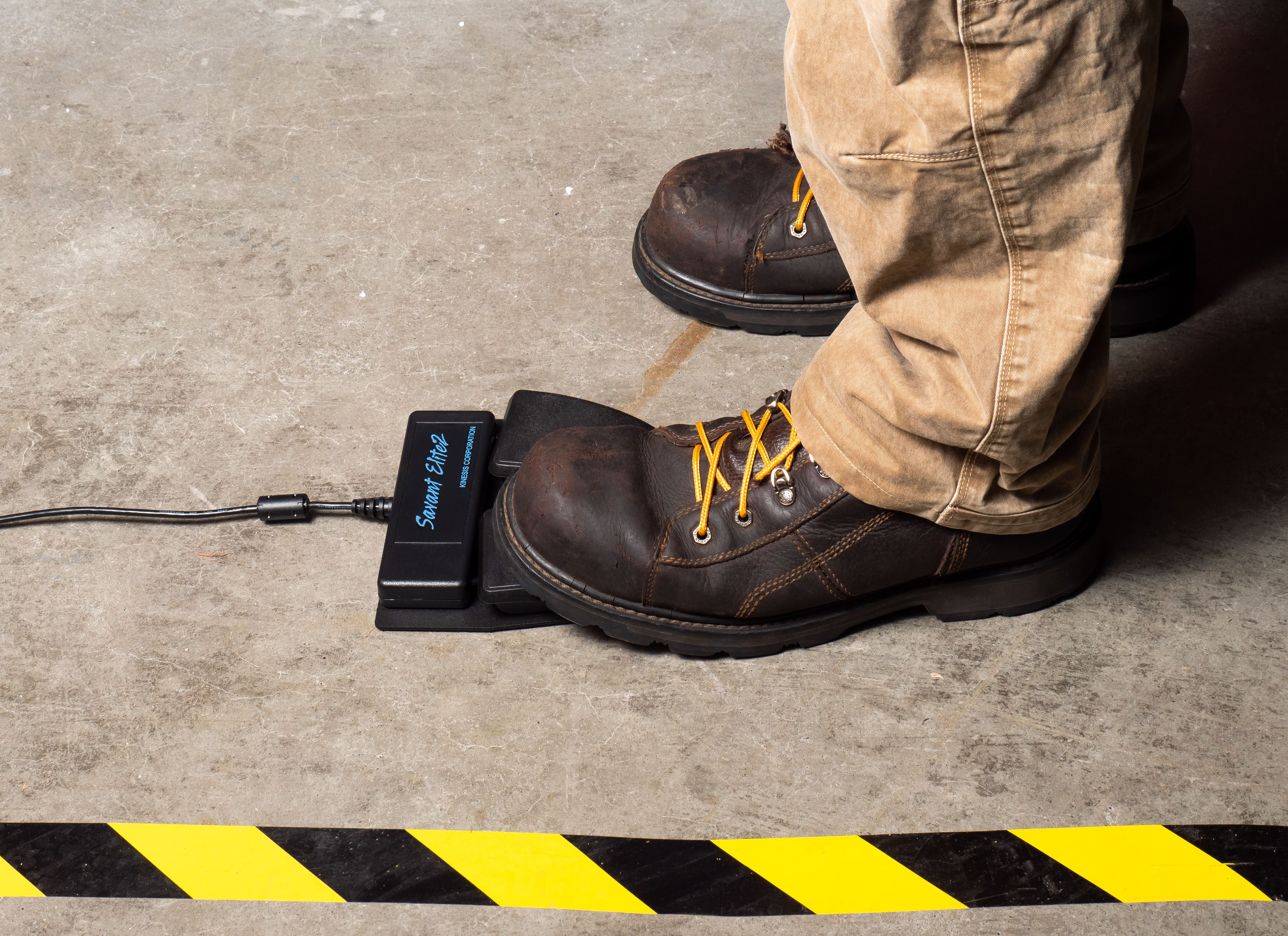 Worker in safety boots stepping on black foot pedal, industrial floor with yellow hazard stripe in foreground.