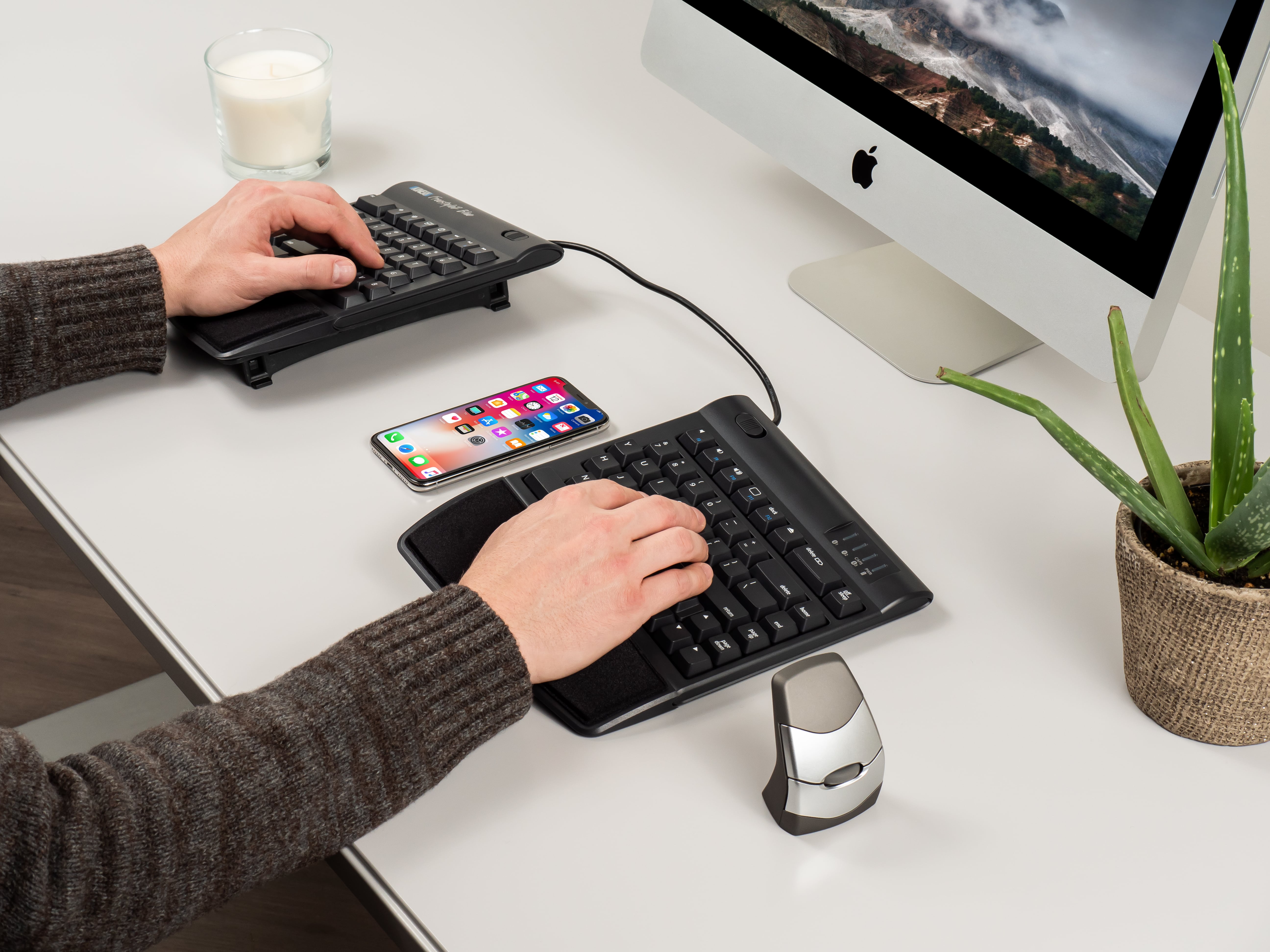 Ergonomic dual keyboard setup with a computer and smartphone on a desk, featuring a candle and aloe plant for aesthetics.