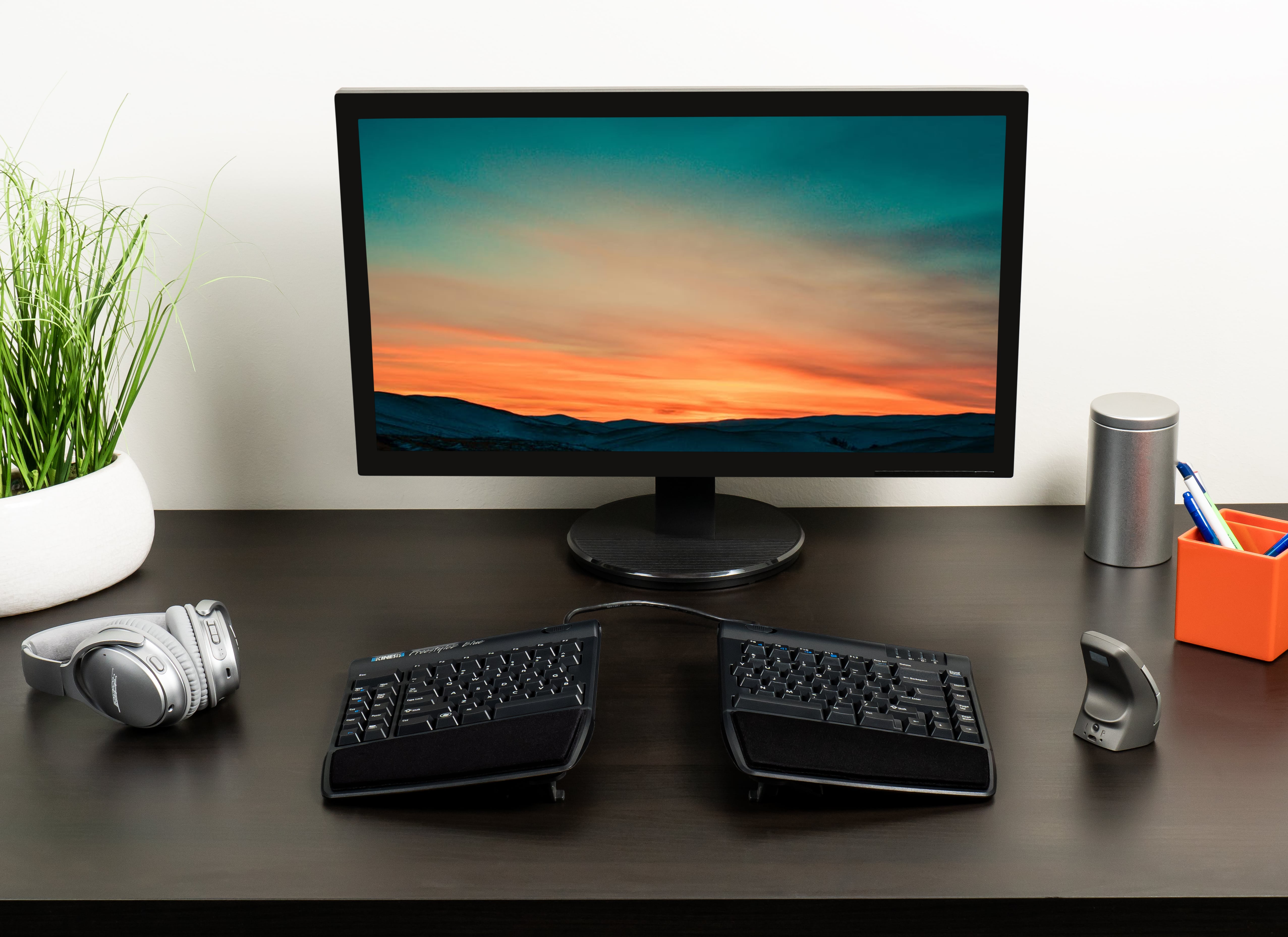 Ergonomic computer setup with split keyboard, vertical mouse, headphones, and sunset screen on a modern desk.