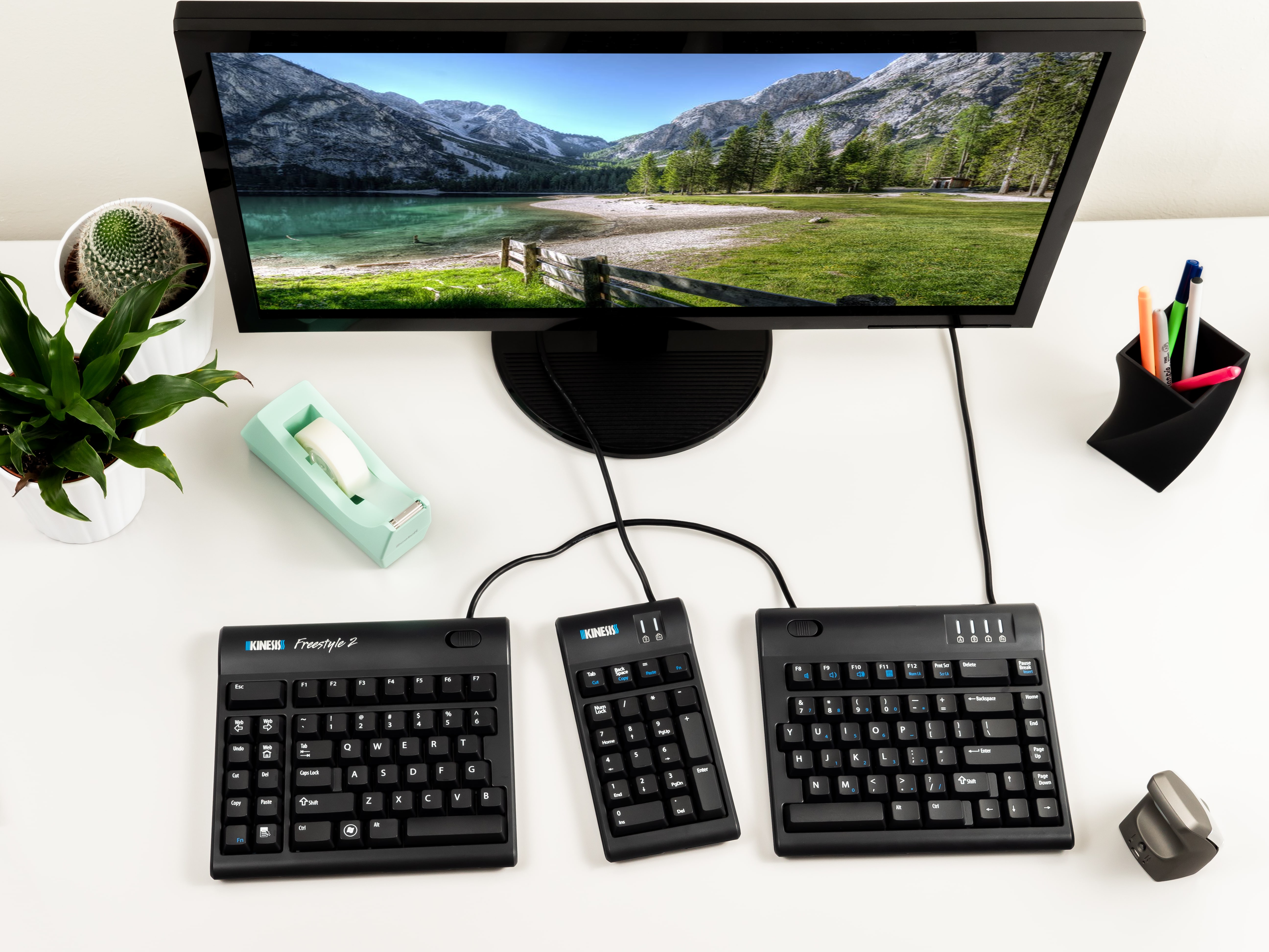 Ergonomic computer setup with split keyboard on desk, monitor displaying scenic mountain view, plants, and office supplies.