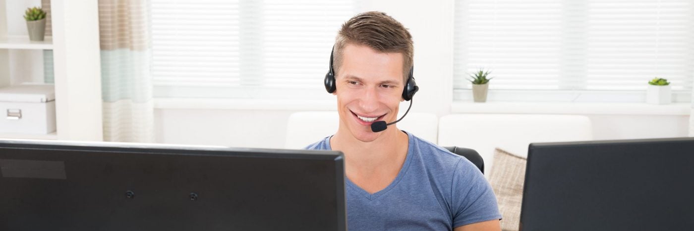 Smiling customer service representative wearing headset at desk, working on multiple monitors.