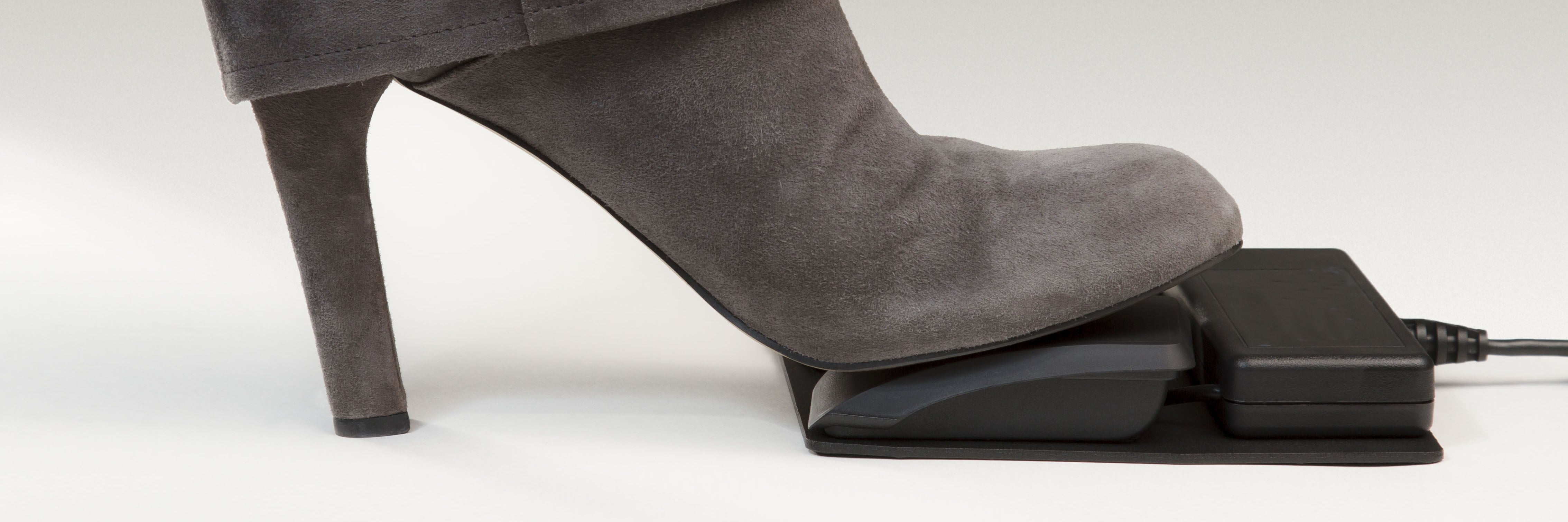 Women's high-heeled boot pressing a black foot pedal on a light background.