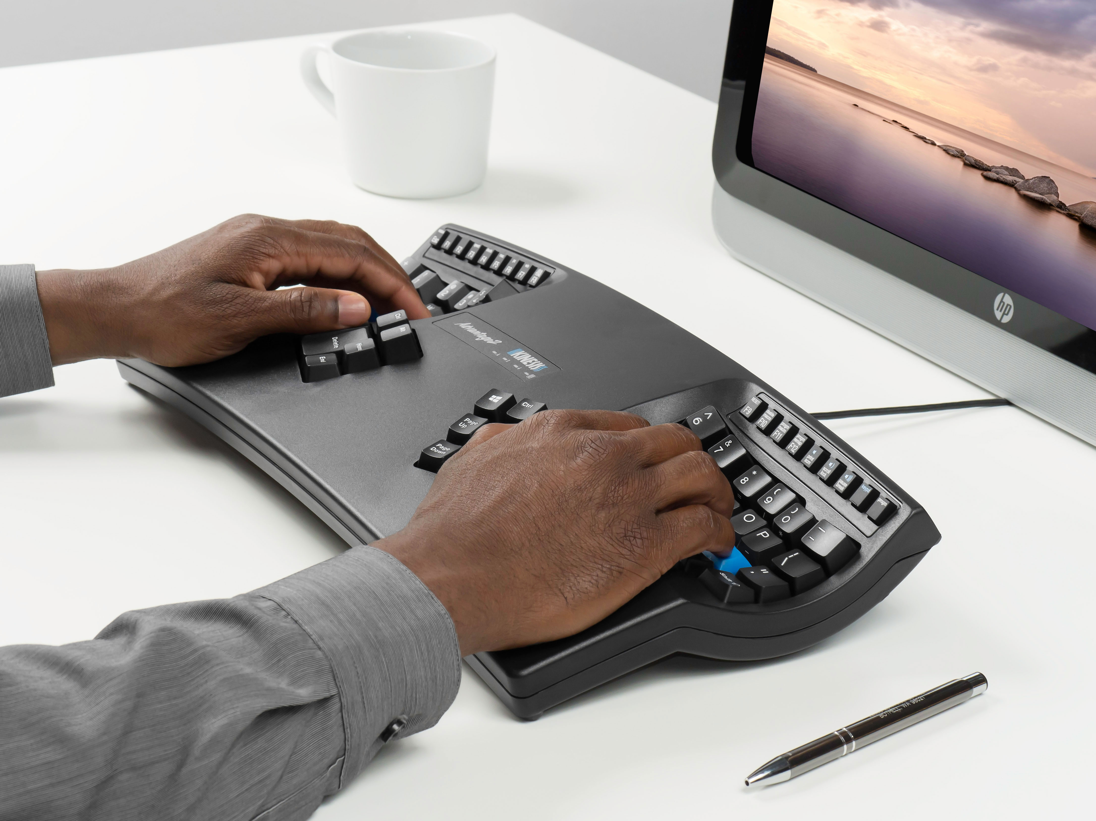 Person typing on an ergonomic keyboard at a desk with a computer, coffee mug, and pen nearby.