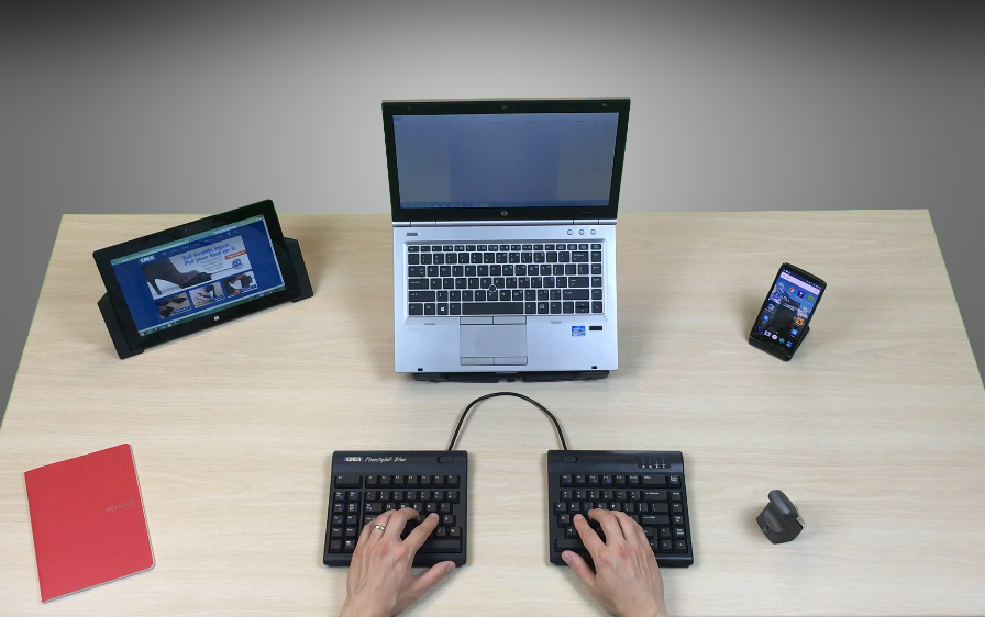 Person typing on a split keyboard connected to a laptop, with a tablet and phone on a desk.