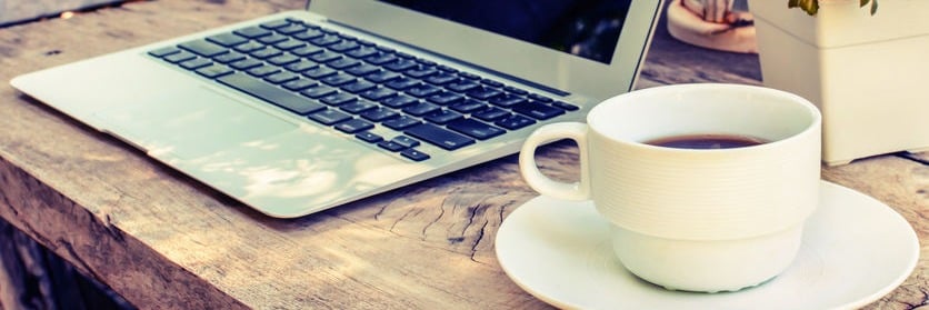 Laptop on wooden table with coffee cup; cozy workspace outdoors.