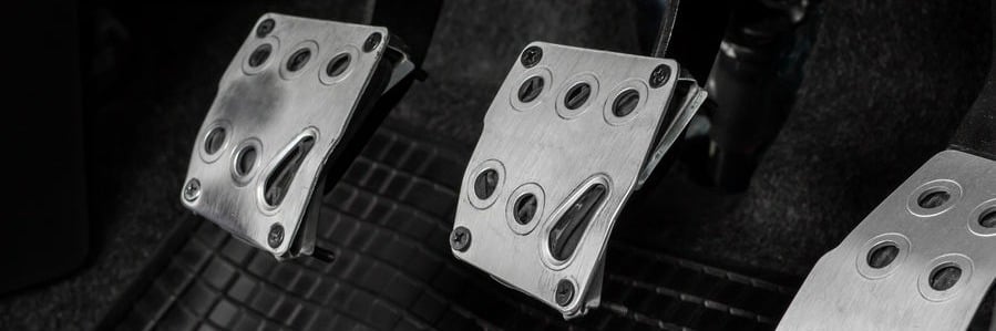 Close-up of metal car pedals: accelerator, brake, and clutch for a manual transmission, on a textured floor mat.