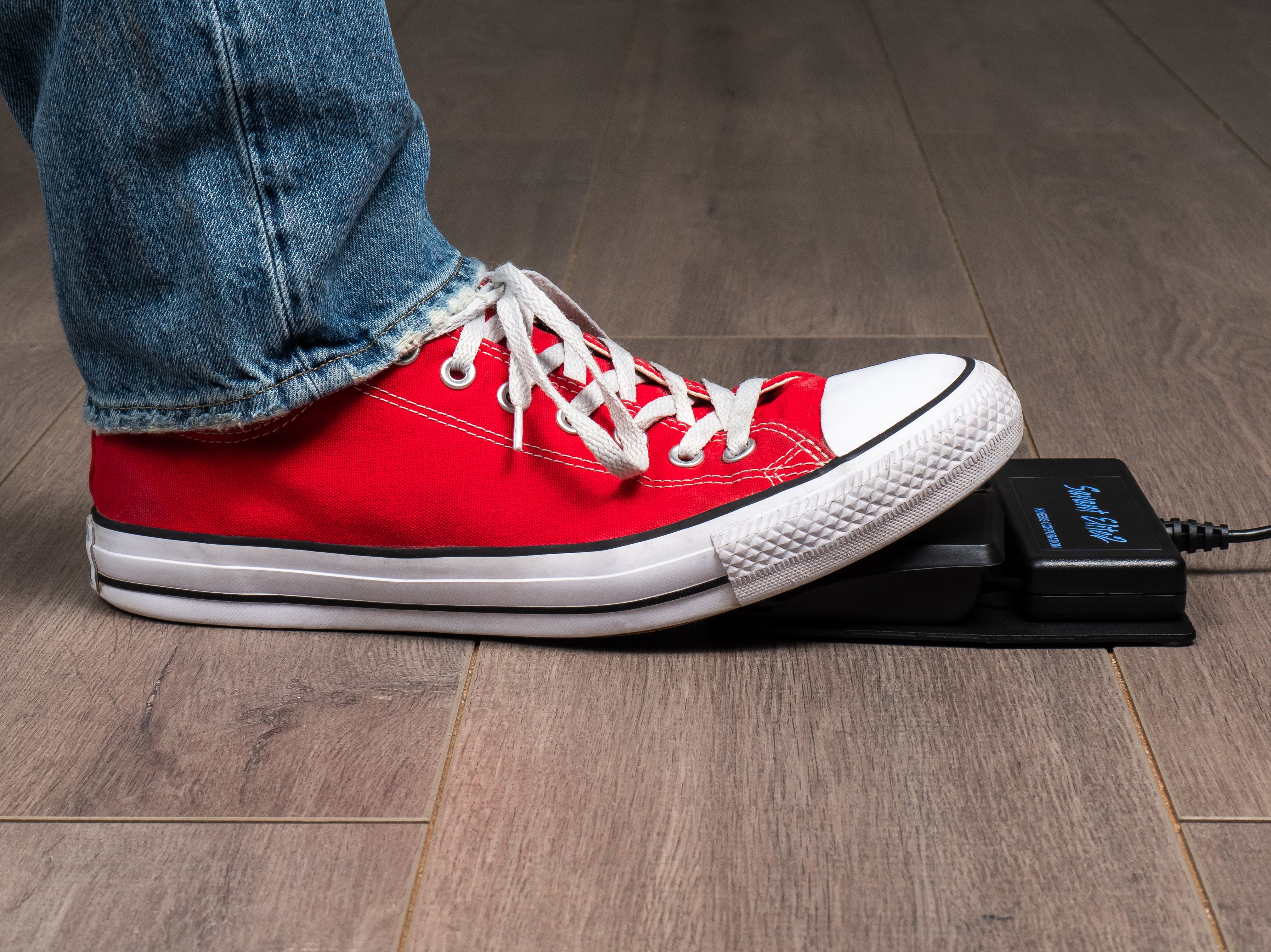 Red sneaker pressing foot pedal on wooden floor, showcasing casual footwear and technology interaction.