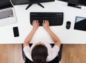 Overhead view of person typing on keyboard at a desk with multiple screens, smartphone, laptop, and tablet.
