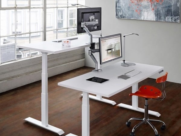 Modern ergonomic office setup with adjustable white desks and computers, featuring a sleek red chair and large window view.